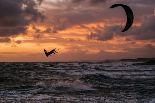 Un kitesurfeur en plein saut très haut au-dessus de l'eau, silhouette et aile détachées sur un ciel de fin de journée, mer agitée en contrebas