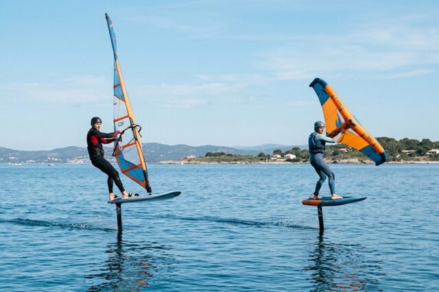 Un windfoiler avec son gréement et un wingfoiler avec son aile à main, tous deux en vol sur le même plan d'eau