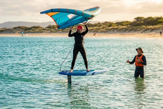 Une pratiquante casquée décolle sur son foil en wingfoil, aile tenue au-dessus de la tête, un moniteur debout dans l'eau à côté d'elle